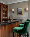 A traditional home bar featuring rich cherry wood cabinetry, a polished brass bar faucet, and a black marble countertop. Walls are painted in a soft greige, and the space is illuminated by classic wall sconces and recessed lighting, with emerald green velvet bar stools. — close-up of textures and materials