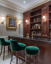 A traditional home bar featuring rich cherry wood cabinetry, a polished brass bar faucet, and a black marble countertop. Walls are painted in a soft greige, and the space is illuminated by classic wall sconces and recessed lighting, with emerald green velvet bar stools. — styling and décor detail