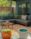 A family-friendly Mid-Century Modern patio featuring a low-profile charcoal grey L-shaped sectional with olive green performance fabric cushions, a geometric outdoor rug in burnt orange and teal, and teak wood accent tables. Large, sculptural ceramic planters with pet-safe plants are arranged around the seating area, with a subtle concrete water bowl for pets integrated into the design.