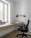 Modern home office with a minimalist aesthetic featuring a light oak floating desk, a black ergonomic chair, white walls, and a sleek matte black desk lamp. — alternate angle