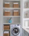 Small coastal laundry room featuring white shiplap walls, a stackable washer and dryer, open shelving with woven baskets, and a light quartz countertop with a small farmhouse sink. Soft blue accents are visible in a decorative ceramic vase.