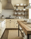 A cozy rustic kitchen featuring creamy off-white cabinets, a reclaimed wood island, and open shelving displaying white stoneware. Wrought iron hardware and a sisal runner add to the warm, inviting atmosphere, with natural light streaming in.