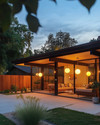 A Mid-Century Modern exterior at dusk, featuring a low-slung roofline, large glass windows, and warm glow from spherical globe sconces flanking the entry. A Sputnik chandelier is visible under a covered patio, illuminating a seating area with clean-lined furniture and a natural wood fence.
