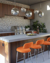A stylish Mid-Century Modern outdoor kitchen featuring a grey terrazzo countertop, rich walnut-toned cabinetry, and a sleek brushed stainless steel grill. Orange upholstered bar stools with chrome legs sit at the island, under a cluster of spherical globe pendant lights. — close-up of textures and materials