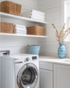 Small coastal laundry room featuring white shiplap walls, a stackable washer and dryer, open shelving with woven baskets, and a light quartz countertop with a small farmhouse sink. Soft blue accents are visible in a decorative ceramic vase. — close-up of textures and materials