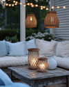 A coastal patio at dusk with warm string lights strung overhead, rattan pendant lights, and hurricane lanterns on a weathered wood table, surrounded by comfortable white cushions and soft blue throws. — close-up of textures and materials