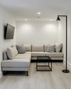 A small modern basement living area featuring a light gray modular sectional sofa, a black metal coffee table, and a wall-mounted television. White walls and light wood flooring contribute to an open, airy feel, complemented by recessed lighting and a tall, slender floor lamp. — close-up of textures and materials
