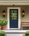 A cozy traditional exterior featuring a warm gray facade with creamy white trim, a deep blue front door, and classic black lantern sconces flanking the entrance. Potted green plants in terracotta pots sit on either side of the door, and a traditional wooden bench offers a welcoming touch.