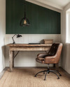 A cozy rustic home office featuring a reclaimed oak desk, a distressed cognac leather chair, and a jute rug. Walls are painted a warm off-white, complemented by a deep forest green accent wall, with an iron pendant light illuminating the space. — alternate angle