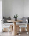 A small, bright Scandinavian breakfast nook featuring a light wood pedestal table, two minimalist white chairs, and a built-in banquette with grey linen cushions. A small potted plant sits on the table, and white walls create an airy, open feel. — styling and décor detail