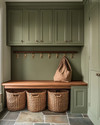 A traditional mudroom with sage green built-in cabinetry, a natural wood bench, and flagstone flooring. Woven rattan baskets are neatly tucked under the bench, and aged brass hooks line the wall. — close-up of textures and materials