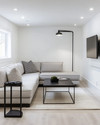 A small modern basement living area featuring a light gray modular sectional sofa, a black metal coffee table, and a wall-mounted television. White walls and light wood flooring contribute to an open, airy feel, complemented by recessed lighting and a tall, slender floor lamp. — alternate angle