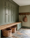 A traditional mudroom with sage green built-in cabinetry, a natural wood bench, and flagstone flooring. Woven rattan baskets are neatly tucked under the bench, and aged brass hooks line the wall.