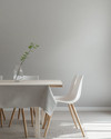 A bright Scandinavian dining room featuring a light birch wood dining table, minimalist white chairs, a simple linen tablecloth, and a clear glass vase with a single green branch, all set against soft grey walls.