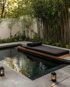 A serene Japandi pool area featuring a dark-tiled swimming pool surrounded by smooth concrete pavers and a low-slung teak daybed with charcoal cushions. Subtle solar-powered lanterns glow amidst pet-friendly green groundcover and slender bamboo plants. — styling and décor detail