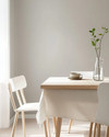 A bright Scandinavian dining room featuring a light birch wood dining table, minimalist white chairs, a simple linen tablecloth, and a clear glass vase with a single green branch, all set against soft grey walls. — styling and décor detail