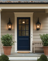 A cozy traditional exterior featuring a warm gray facade with creamy white trim, a deep blue front door, and classic black lantern sconces flanking the entrance. Potted green plants in terracotta pots sit on either side of the door, and a traditional wooden bench offers a welcoming touch. — alternate angle