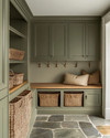 A traditional mudroom with sage green built-in cabinetry, a natural wood bench, and flagstone flooring. Woven rattan baskets are neatly tucked under the bench, and aged brass hooks line the wall. — alternate angle