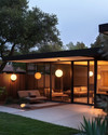 A Mid-Century Modern exterior at dusk, featuring a low-slung roofline, large glass windows, and warm glow from spherical globe sconces flanking the entry. A Sputnik chandelier is visible under a covered patio, illuminating a seating area with clean-lined furniture and a natural wood fence. — styling and décor detail