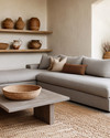 A Modern Craft living room with a large, comfortable sectional sofa in a warm grey performance fabric, an oak coffee table with a matte finish, and a woven jute rug. Walls are painted in a soft off-white, complemented by shelves displaying hand-thrown ceramic vases and a large woven basket for storage. — alternate angle