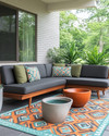 A family-friendly Mid-Century Modern patio featuring a low-profile charcoal grey L-shaped sectional with olive green performance fabric cushions, a geometric outdoor rug in burnt orange and teal, and teak wood accent tables. Large, sculptural ceramic planters with pet-safe plants are arranged around the seating area, with a subtle concrete water bowl for pets integrated into the design. — alternate angle