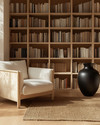 A Japandi library featuring light blonde wood bookshelves filled with books, a comfortable cream linen armchair, and a natural jute rug on light hardwood floors, accented by a matte black ceramic vase. — close-up of textures and materials