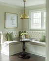 A cozy traditional breakfast nook featuring a built-in cream banquette with sage green cushions, a round dark wood pedestal table, and a classic brass pendant light, all bathed in soft morning light.