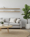 A bright Scandinavian family room featuring a light grey sofa with linen cushions, a round blonde wood coffee table, and a large jute rug. Walls are painted in a soft off-white, complemented by a tall fiddle leaf fig plant in a white pot, and minimalist floating shelves. — alternate angle