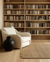 A Japandi library featuring light blonde wood bookshelves filled with books, a comfortable cream linen armchair, and a natural jute rug on light hardwood floors, accented by a matte black ceramic vase. — alternate angle