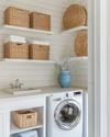 Small coastal laundry room featuring white shiplap walls, a stackable washer and dryer, open shelving with woven baskets, and a light quartz countertop with a small farmhouse sink. Soft blue accents are visible in a decorative ceramic vase. — alternate angle