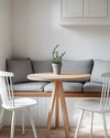 A small, bright Scandinavian breakfast nook featuring a light wood pedestal table, two minimalist white chairs, and a built-in banquette with grey linen cushions. A small potted plant sits on the table, and white walls create an airy, open feel. — alternate angle