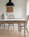 A bright farmhouse dining room featuring a distressed oak dining table, spindle-back chairs, and a woven rug. A large, vintage-inspired matte black pendant light hangs above, with a galvanized metal pitcher and white linen runner on the table. Walls are painted in a creamy off-white.