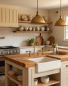 A cozy farmhouse kitchen features creamy off-white shaker cabinets, a large oak butcher block island, and a ceramic farmhouse sink. Warm brass pendant lights hang above the island, illuminating open shelves displaying vintage ceramic dishes and cookbooks.