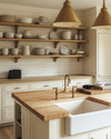 A cozy farmhouse kitchen features creamy off-white shaker cabinets, a large oak butcher block island, and a ceramic farmhouse sink. Warm brass pendant lights hang above the island, illuminating open shelves displaying vintage ceramic dishes and cookbooks. — styling and décor detail