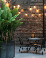 Industrial exterior with galvanized steel planters holding tall green plants, a black metal bistro set with a concrete top, and string lights with exposed Edison bulbs draped over a patio. — view 3