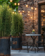 Industrial exterior with galvanized steel planters holding tall green plants, a black metal bistro set with a concrete top, and string lights with exposed Edison bulbs draped over a patio.