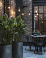Industrial exterior with galvanized steel planters holding tall green plants, a black metal bistro set with a concrete top, and string lights with exposed Edison bulbs draped over a patio. — view 4