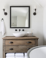 A bright farmhouse bathroom featuring a repurposed distressed wood dresser vanity with a white vessel sink, flanked by matte black barn-style sconces, above which hangs a large mirror with a simple black metal frame. A galvanized metal tub holds rolled white towels. — view 2