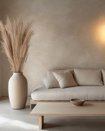 A minimalist Japandi living room featuring a low-profile sofa in natural linen, a light ash wood coffee table, and a large, unglazed ceramic vase with dried pampas grass. The walls are painted in a warm grey, complemented by soft, diffused light from a paper-shade floor lamp. — view 3
