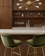 A sleek Mid-Century Modern home bar featuring warm walnut cabinetry, a pristine white quartz countertop, and polished brass bar stools with olive green velvet upholstery. A small Sputnik chandelier hangs above, illuminating a curated collection of glassware and spirits on integrated floating shelves.