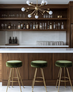 A sleek Mid-Century Modern home bar featuring warm walnut cabinetry, a pristine white quartz countertop, and polished brass bar stools with olive green velvet upholstery. A small Sputnik chandelier hangs above, illuminating a curated collection of glassware and spirits on integrated floating shelves. — view 2