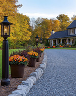 A charming farmhouse driveway leading to a home, featuring a gravel path edged with natural fieldstone, flanked by seasonal planters with colorful mums and pumpkins, and illuminated by dark bronze lantern-style lights on posts. — close-up of textures and materials
