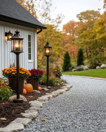 A charming farmhouse driveway leading to a home, featuring a gravel path edged with natural fieldstone, flanked by seasonal planters with colorful mums and pumpkins, and illuminated by dark bronze lantern-style lights on posts. — styling and décor detail