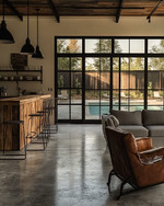 Industrial poolhouse interior featuring polished concrete floors, a reclaimed wood bar, and black steel framed windows looking out to a pool. A charcoal linen sofa, vintage leather armchairs, and matte black industrial pendant lights create a vintage-modern fusion. — close-up of textures and materials