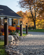 A charming farmhouse driveway leading to a home, featuring a gravel path edged with natural fieldstone, flanked by seasonal planters with colorful mums and pumpkins, and illuminated by dark bronze lantern-style lights on posts. — alternate angle