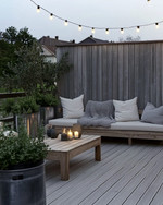 A serene Scandinavian deck with light-toned wooden flooring, a low-profile wooden lounge set with off-white cushions and a chunky knit grey wool throw, minimalist galvanized steel planters with evergreens, and soft string lights overhead. — close-up of textures and materials