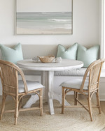 A serene coastal breakfast nook featuring a round bleached oak table with two woven rattan chairs and a light grey upholstered bench, accented with pale seafoam green throw pillows and a natural jute rug. — styling and décor detail