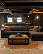 A cozy industrial basement living area with exposed ductwork, a large charcoal sectional sofa, a reclaimed wood coffee table, and an amber-toned area rug, illuminated by warm Edison bulb lighting and task lamps. — styling and décor detail