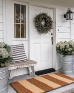 A charming farmhouse exterior with a white front door, layered buffalo check and coir doormats, and a dried eucalyptus wreath. Galvanized metal planters with hydrangeas flank the entrance, beside a distressed wooden bench with a striped throw pillow. — styling and décor detail