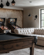 Farmhouse game room with a dark wood billiard table, a cream linen Chesterfield sofa, matte black metal lighting fixtures, and a vintage pinball machine against a wall painted in 'Swiss Coffee'. — styling and décor detail